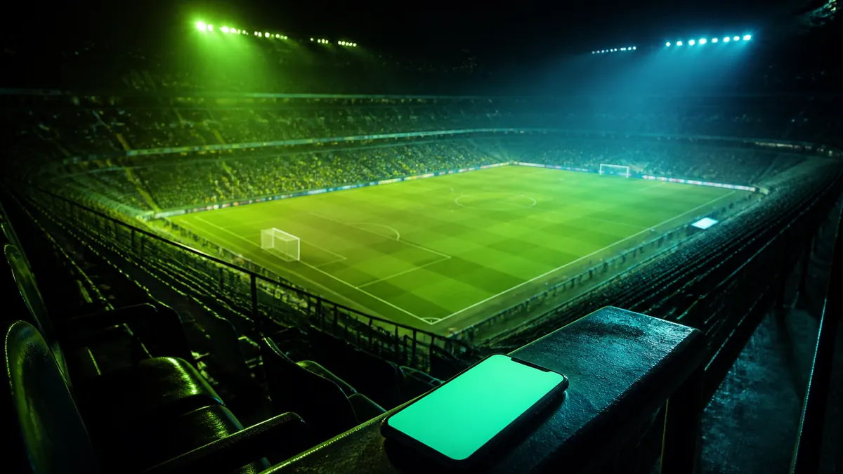 Estadio de fútbol iluminado de noche visto desde la grada con un móvil en primer plano